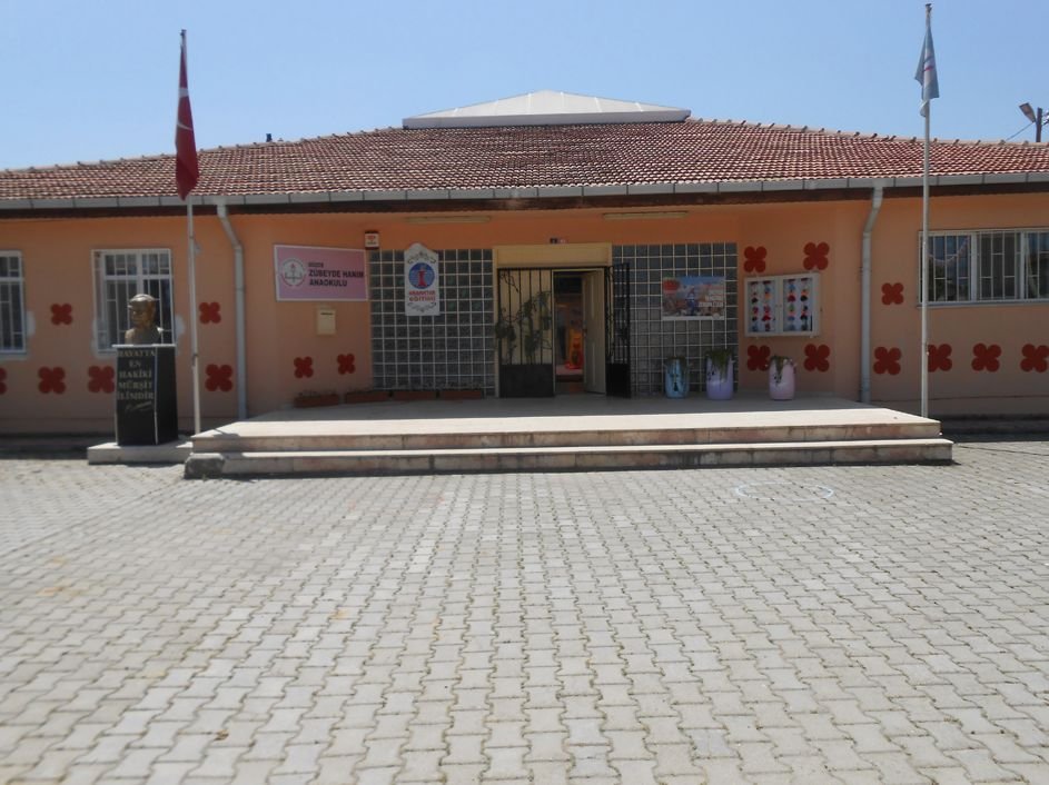 Tek katlı anaokulu binası: kiremit çatı, turuncu cephe, girişte Atatürk büstü ve bayrak direği.